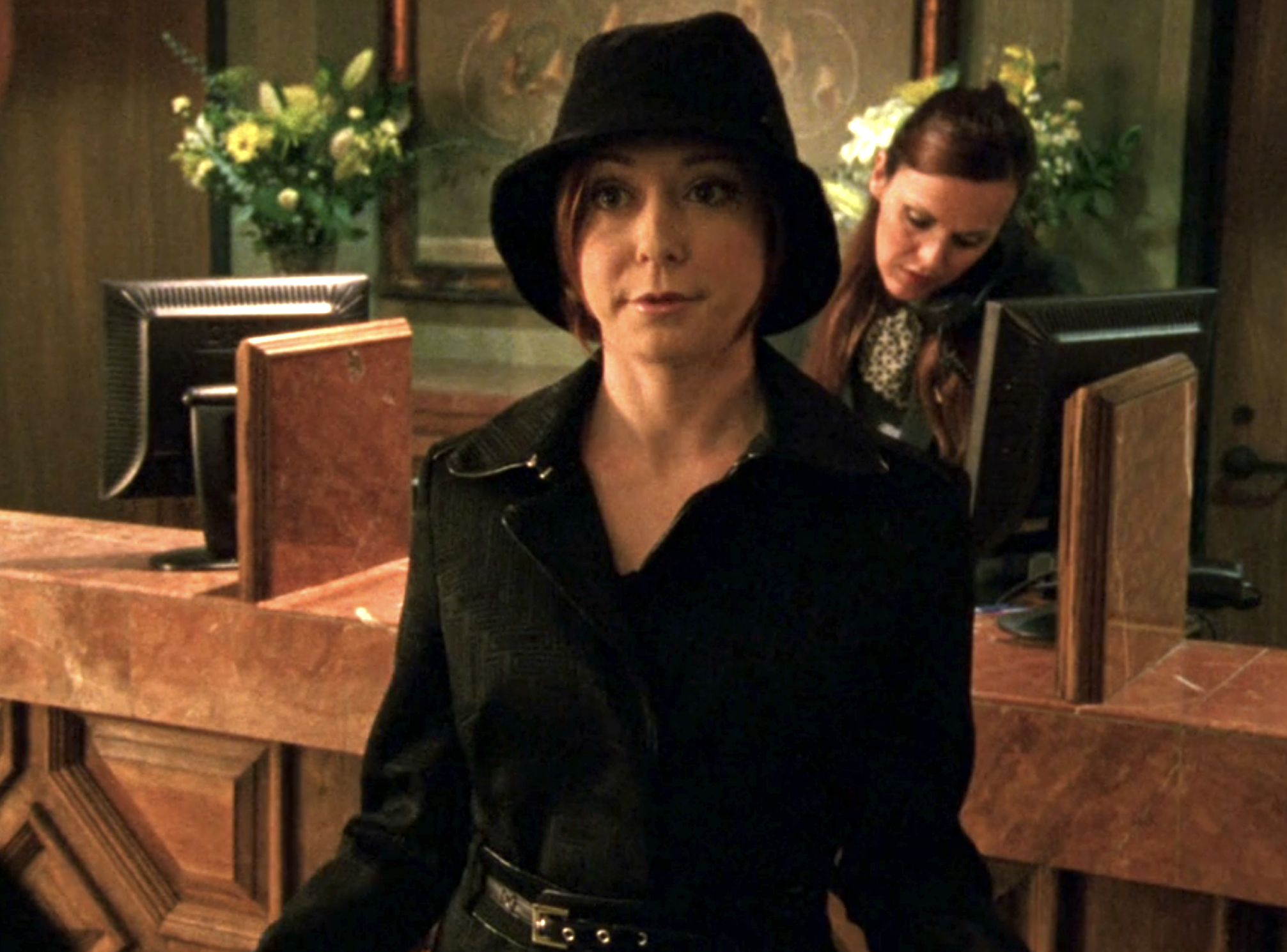 Screenshot from Veronica Mars S1E15. Alyson Hannigan in a black coat and black hat and looking glam is in a hotel lobby.