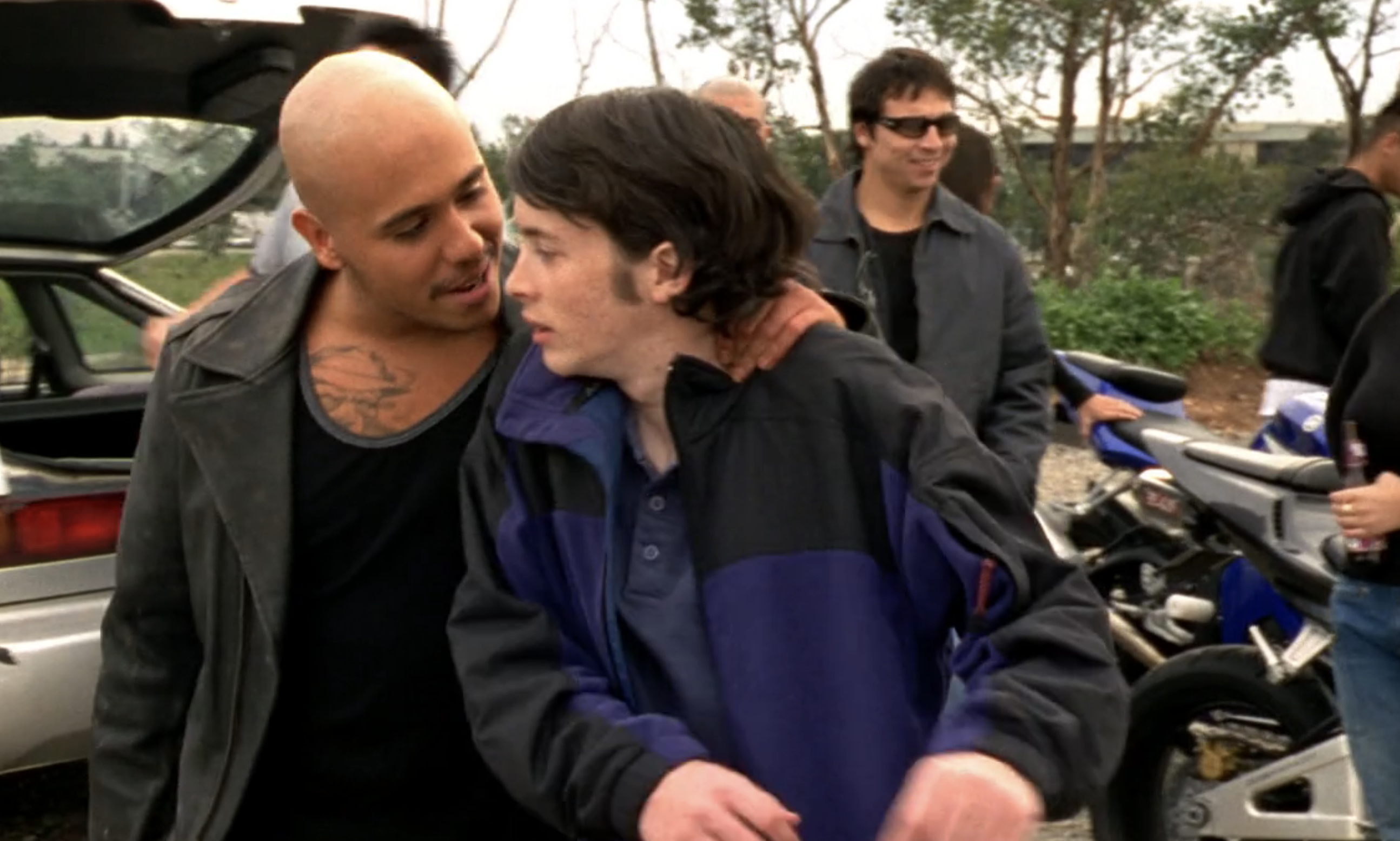 Screenshot from S1E16 of Veronica Mars. Weevil is standing in the foreground with his hand on the scruff of Wilson's neck, looking menacingly at him