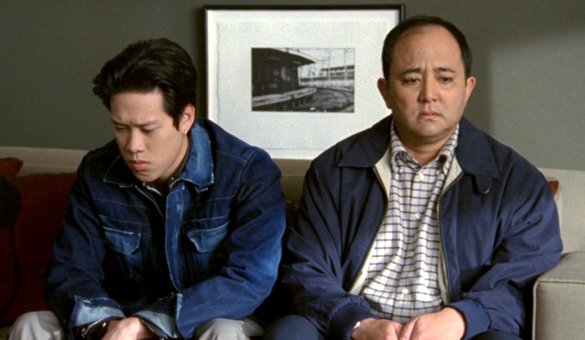 Screenshot from S1E17 of Veronica Mars. Hamilton Cho and his dad sit side by side on a couch. Hamilton is wearing a blue denim jacket. His dad is also weary a navy jacket.