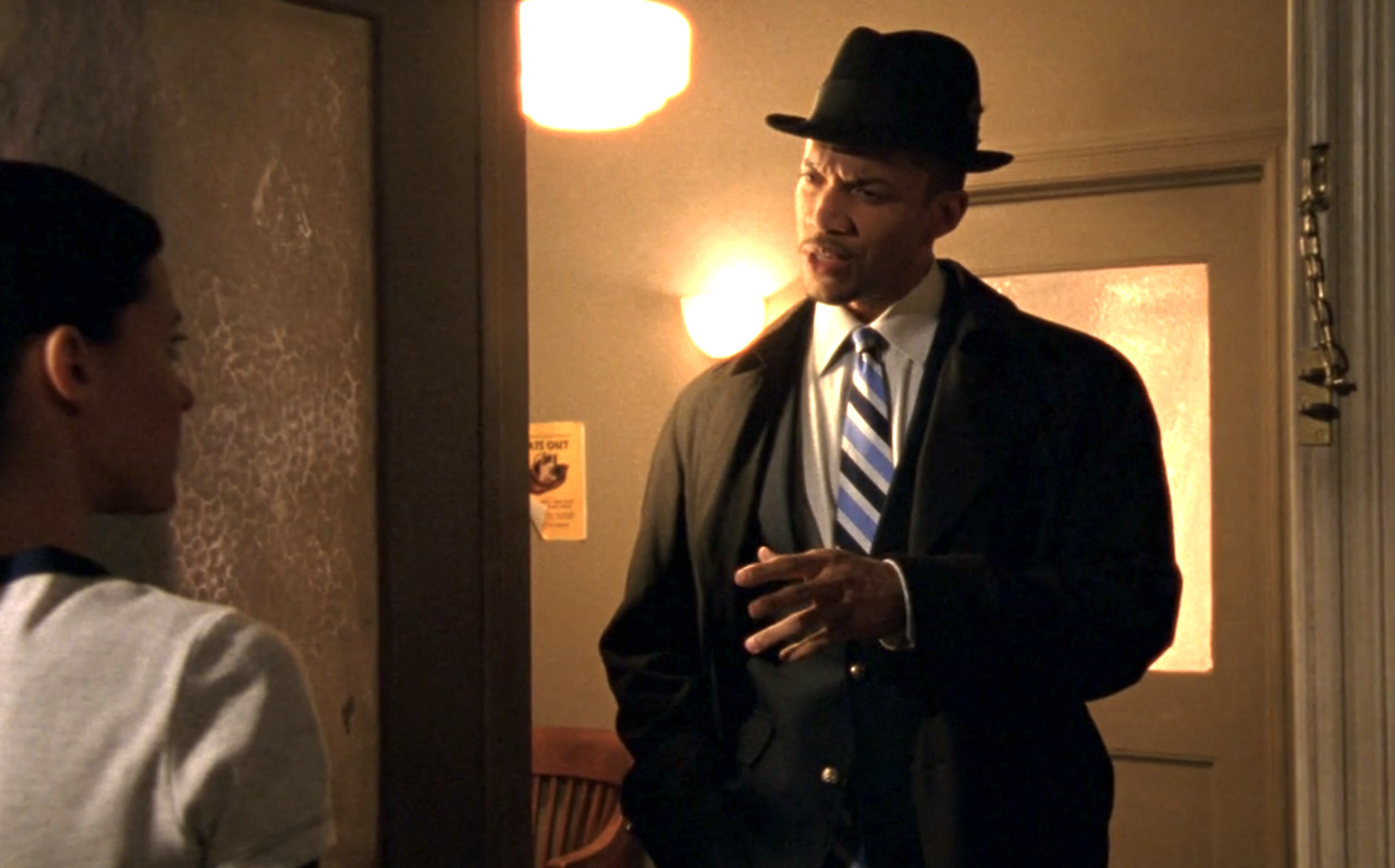 Screenshot from S1E17 of Veronica Mars. Clarence Wiedman is in a doorway talking to a young woman. He's wearing a dark suit, dark coat, and black fedora
