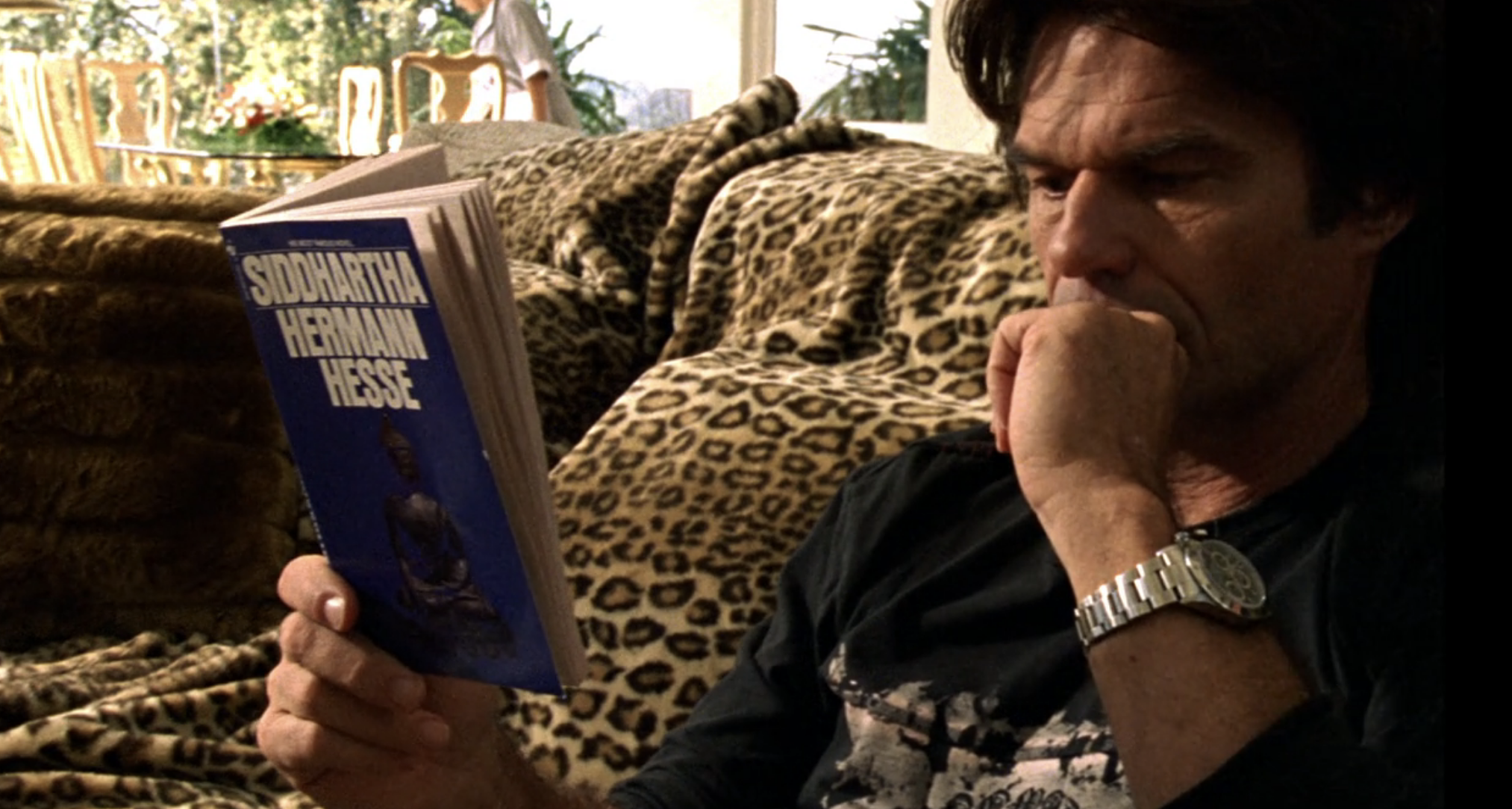 Screenshot from Veronica Mars S1E19. Aaron, a middle aged white man, is seated on a couch which is draped in a leopard print blanket. He's reading Siddhartha by Herman Hess, which he holds in his right hand. His left hand, in a loose fist, rests on his chin.. 