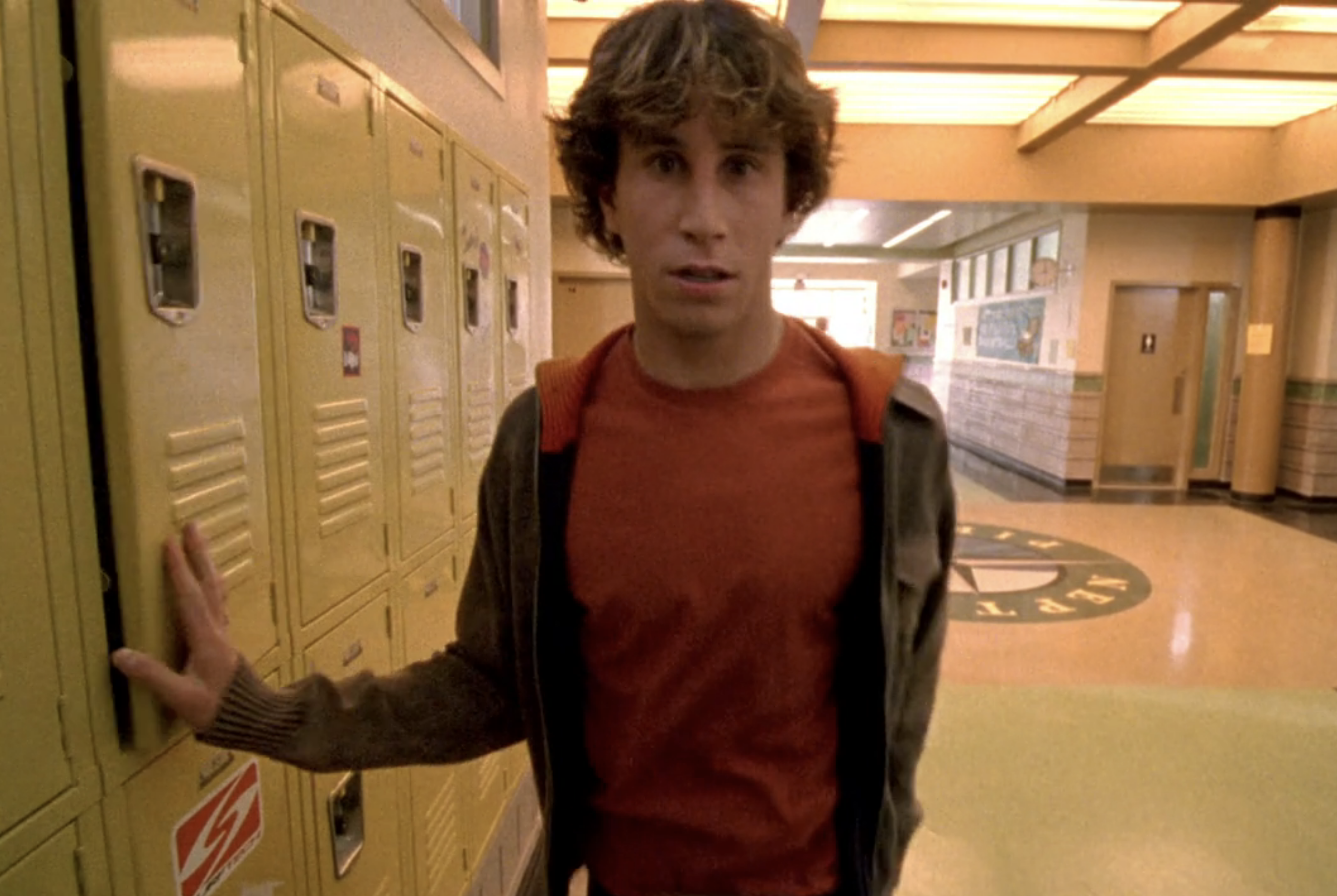 Screenshot from S1E18 of Veronica Mars. Corny is standing in the hallway of Neptune High. His right hand is closing a locker door. He has an expresion of fear on his face.