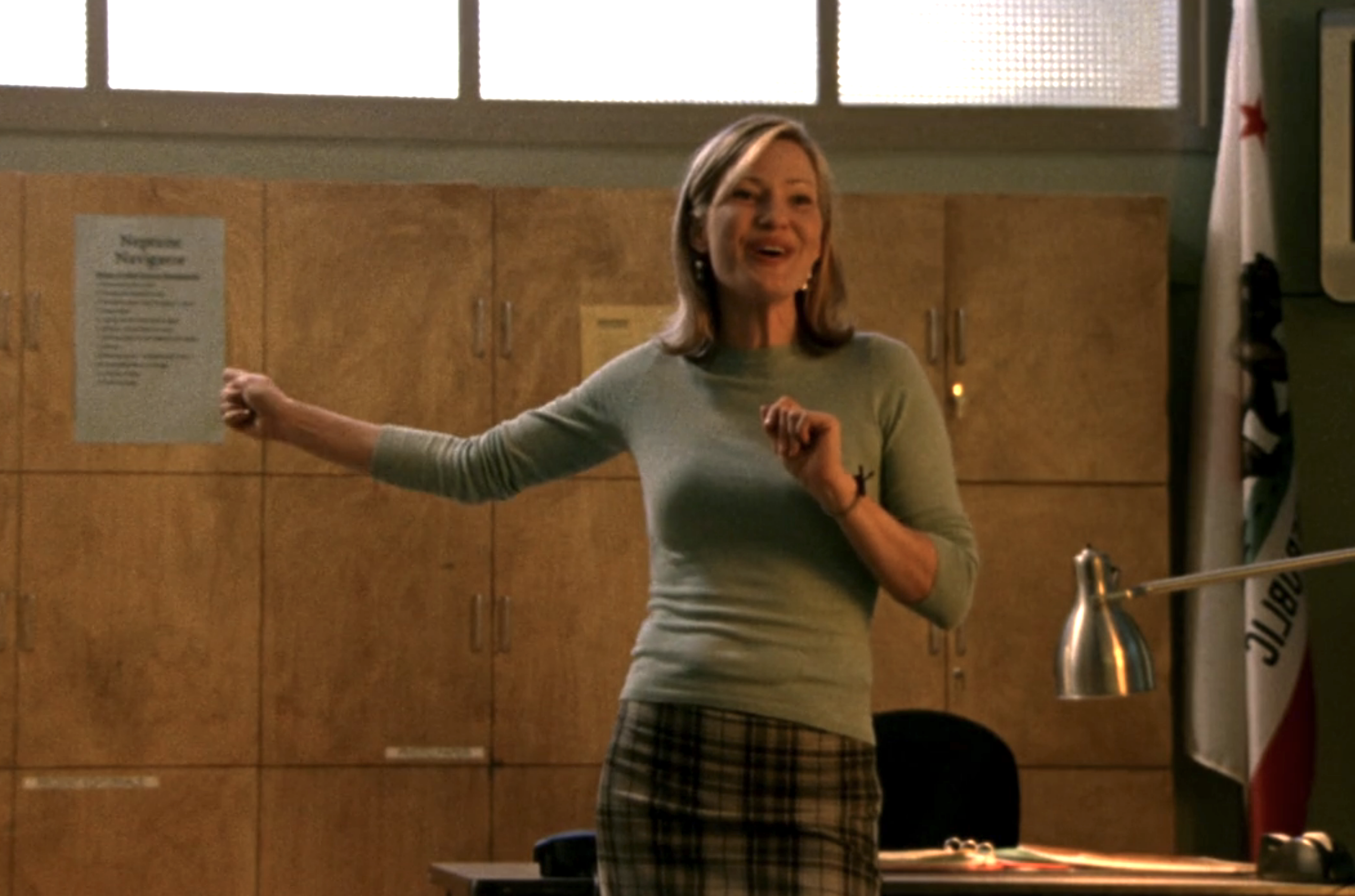 Screenshot from S1E18 of Veronica Mars. Joey Lauren Adams is standing in the front of a classroom smiling and gesturing with her arm.