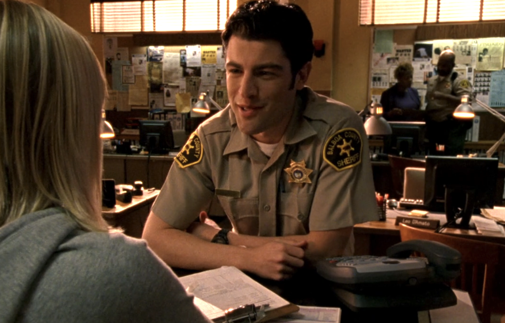 Screenshot from Veronica Mars S1E19. A white man in a sheriff deputy uniform, Leo, is leaning on a counter smiling.