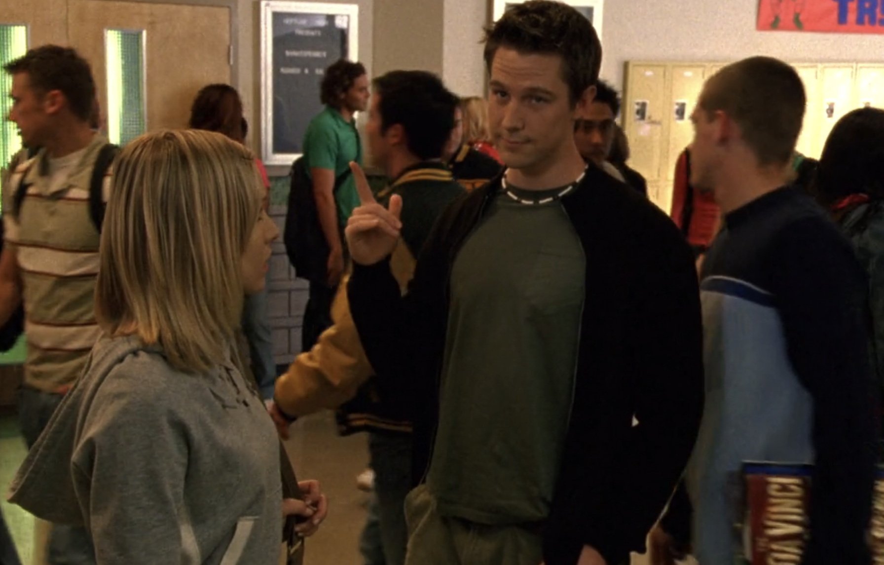 Screenshot from Veronica Mars S1E19. A young white man in a green shirt and dark zip-up sweater is standing in a crowded school hallway. It's Logan and he's pointing up in the air. Veronica is looking at him.