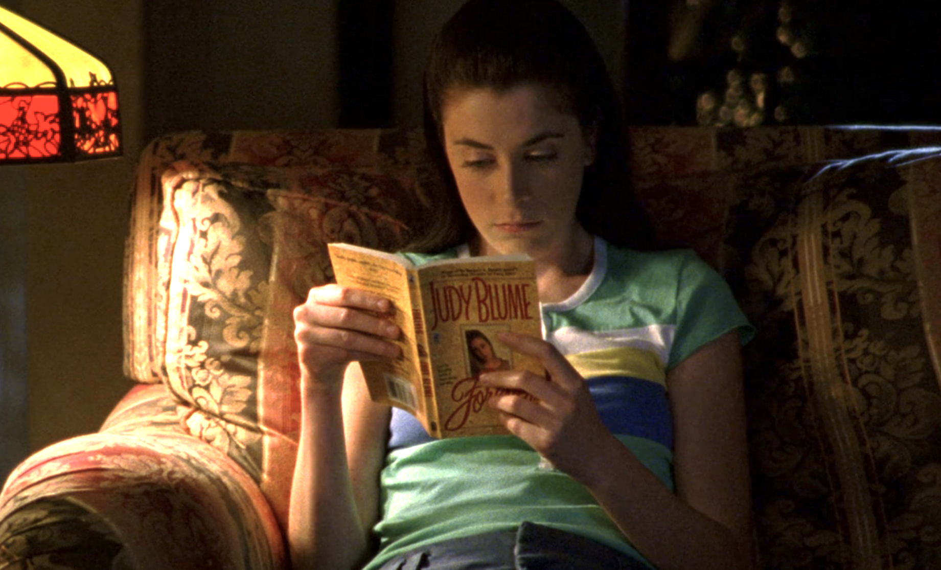 Screenshot from Veronica Mars S1E19. Mandy, young white woman with long brown hair and a green t-shirt, is on a couch reading by lamplight. The book is Forever by Judy Blume.