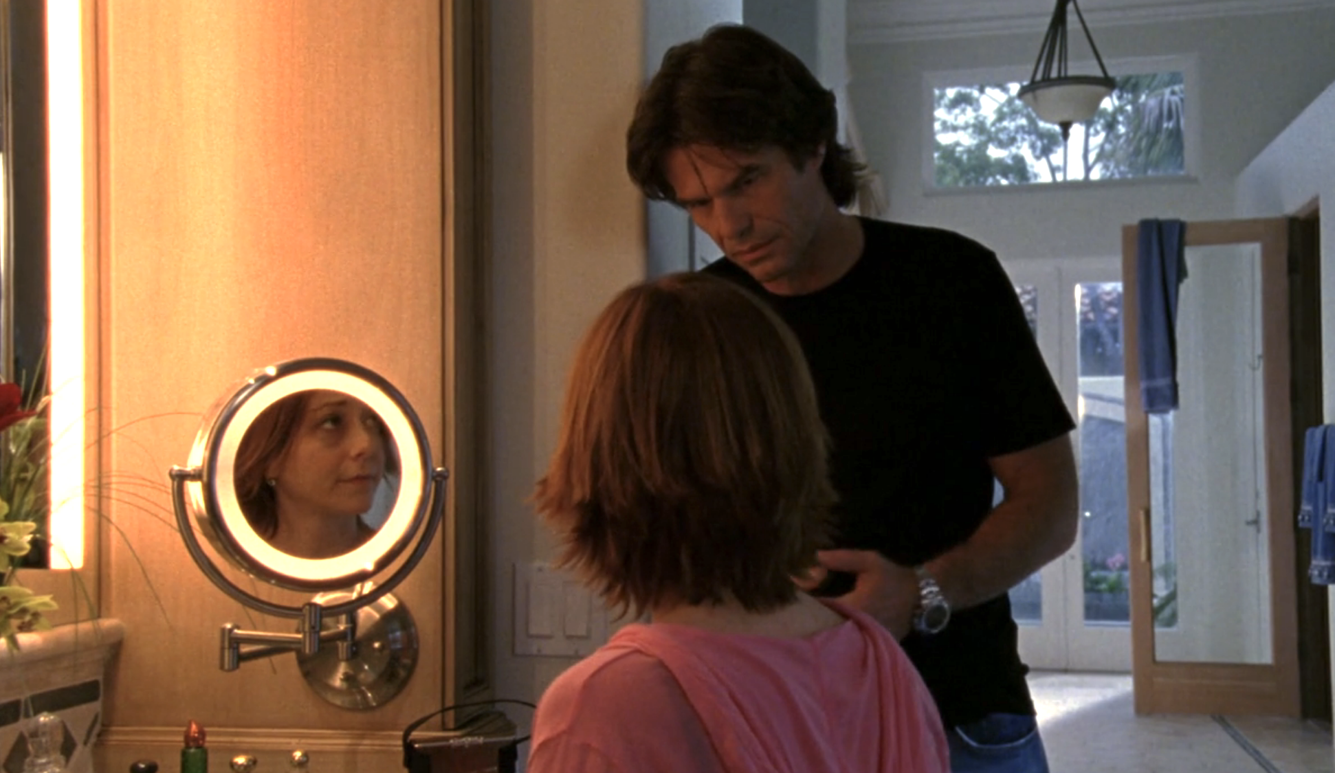 Screenshot from Veronica Mars S1E19. A young white woman, Trina, in a pink hoodie is sitting at a mirror, her back to the camera. We can see her face in the mirror. She has one black eye. Her father Aaron, a middle aged white man in a black tee is standing over her, looking concerned. 