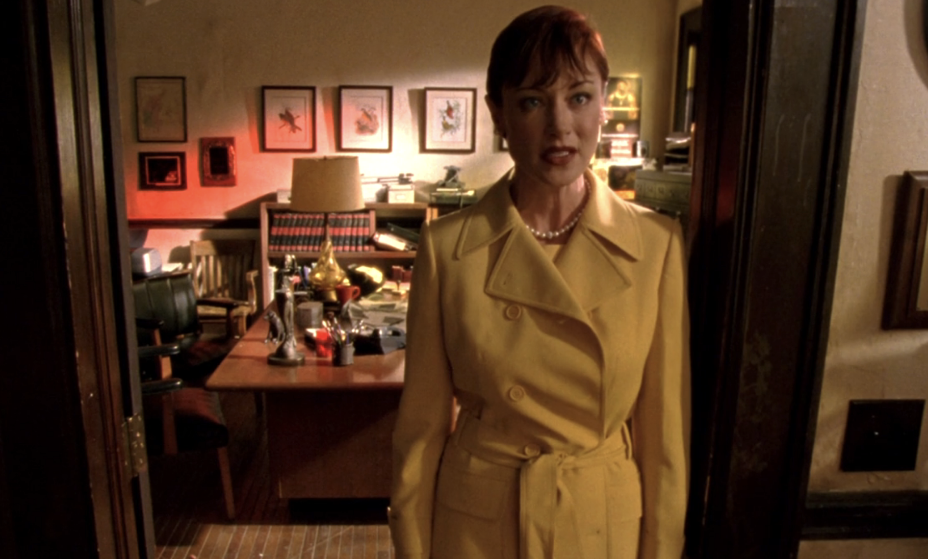 Screenshot from Veronica Mars S1E19. A white woman with a pearl necklace and yellow trenchcoat sants in the doorway of Mars Investigations looking serious.