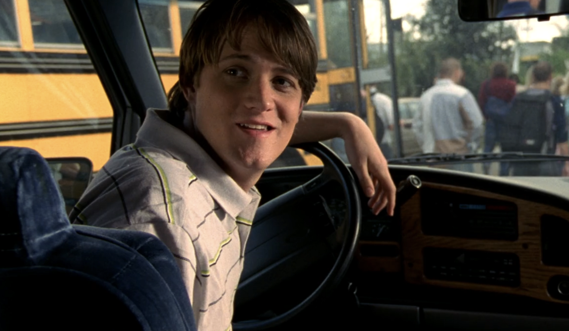 Screenshot from Veronica Mars S1E19. A young white man is in the driver's seat, his left arm resting on the steering wheel. He is turning toward the back of the van and has a smirk on his face.