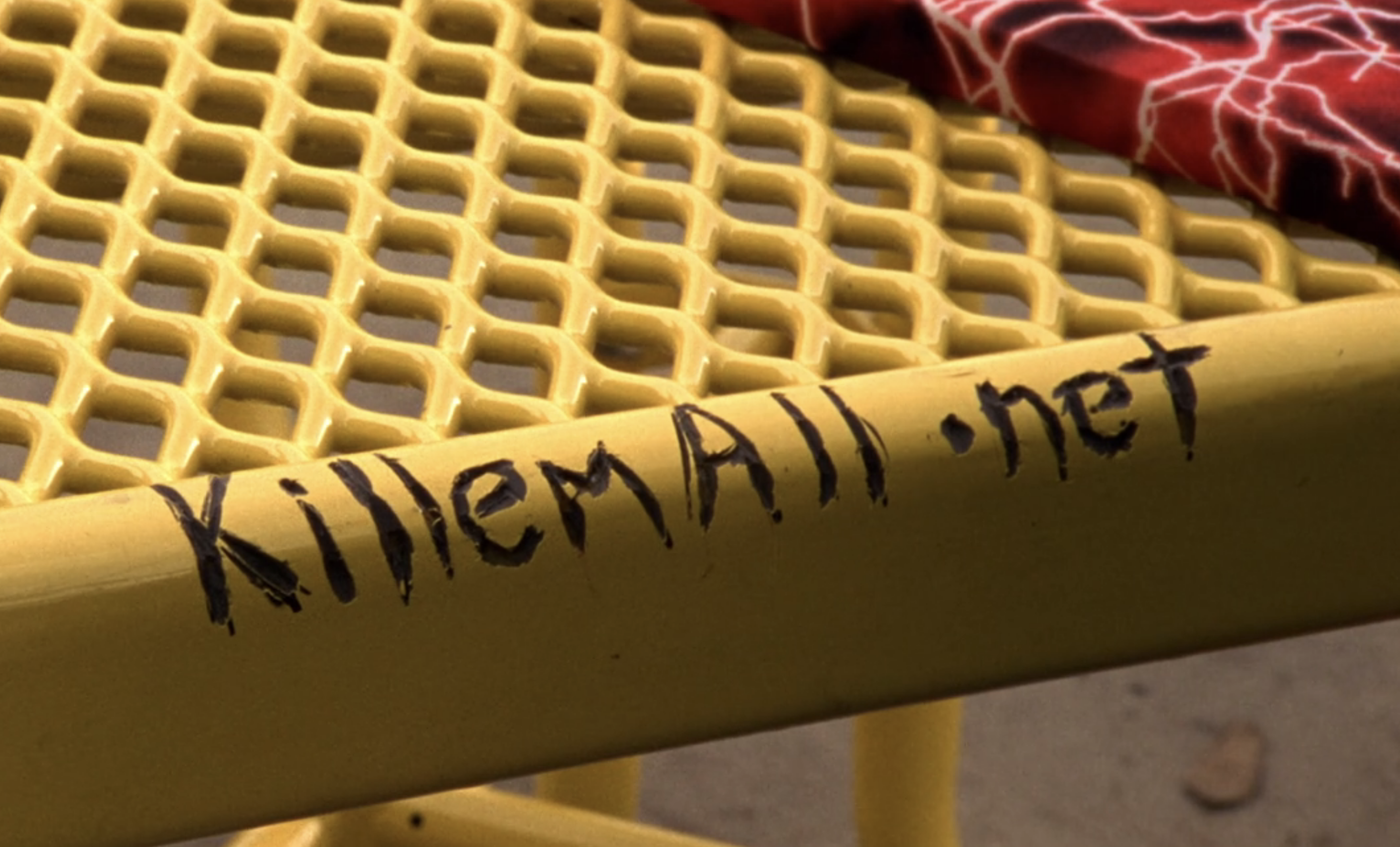 Screenshot from S1E18 of Veronica Mars. A yellow metal table. The words "killemall.net" are scraped into the surface.