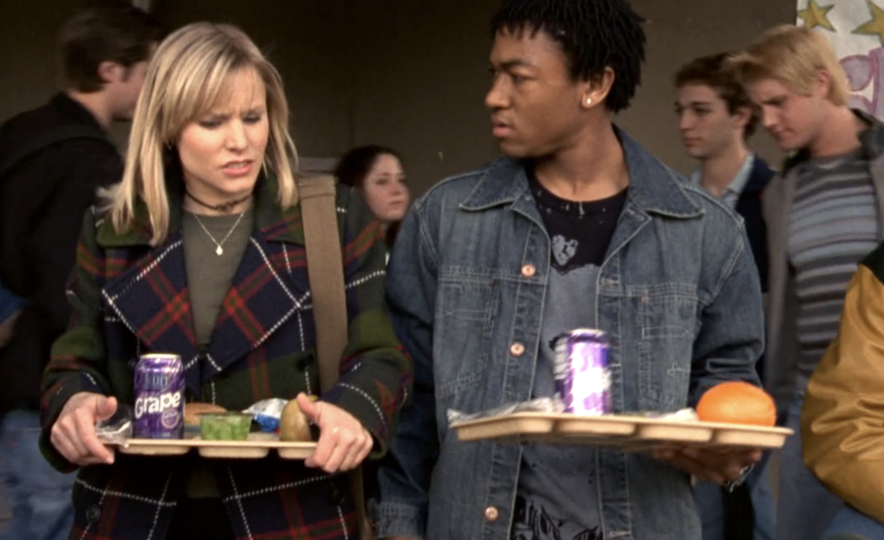 Screenshot from Veronica Mars S1E19. Veronica, a young white woman in a plaid jacket, and Wallace, a young Black man in a jean jacket are walking while carrying lunch trays. They are chatting and both look upset.