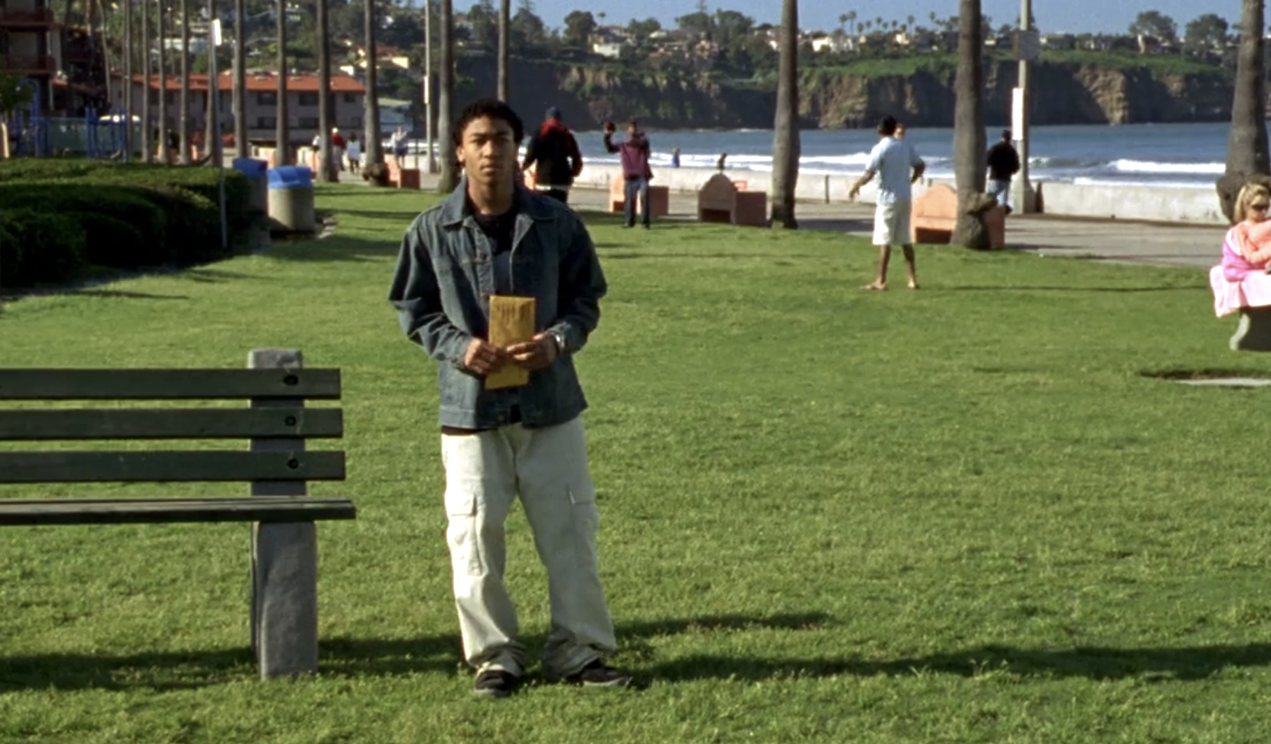 Screenshot from Veronica Mars S1E19. Wallace, a young Black man, is standing on the grass in a park beside a bench. He's wearing a jean jacket and off-white cargo pants and holding an envelope.