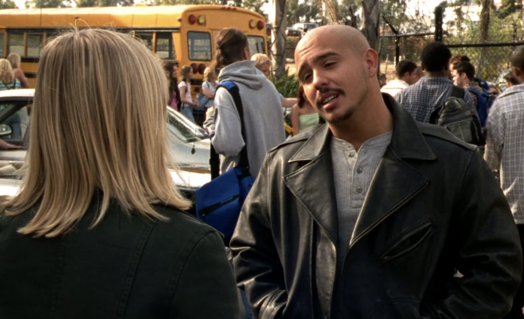 Screenshot from Veronica Mars S1E19. A young Latinx mad in a leather jacket, Weevil, is standing outisde school talkin to Veronica. 