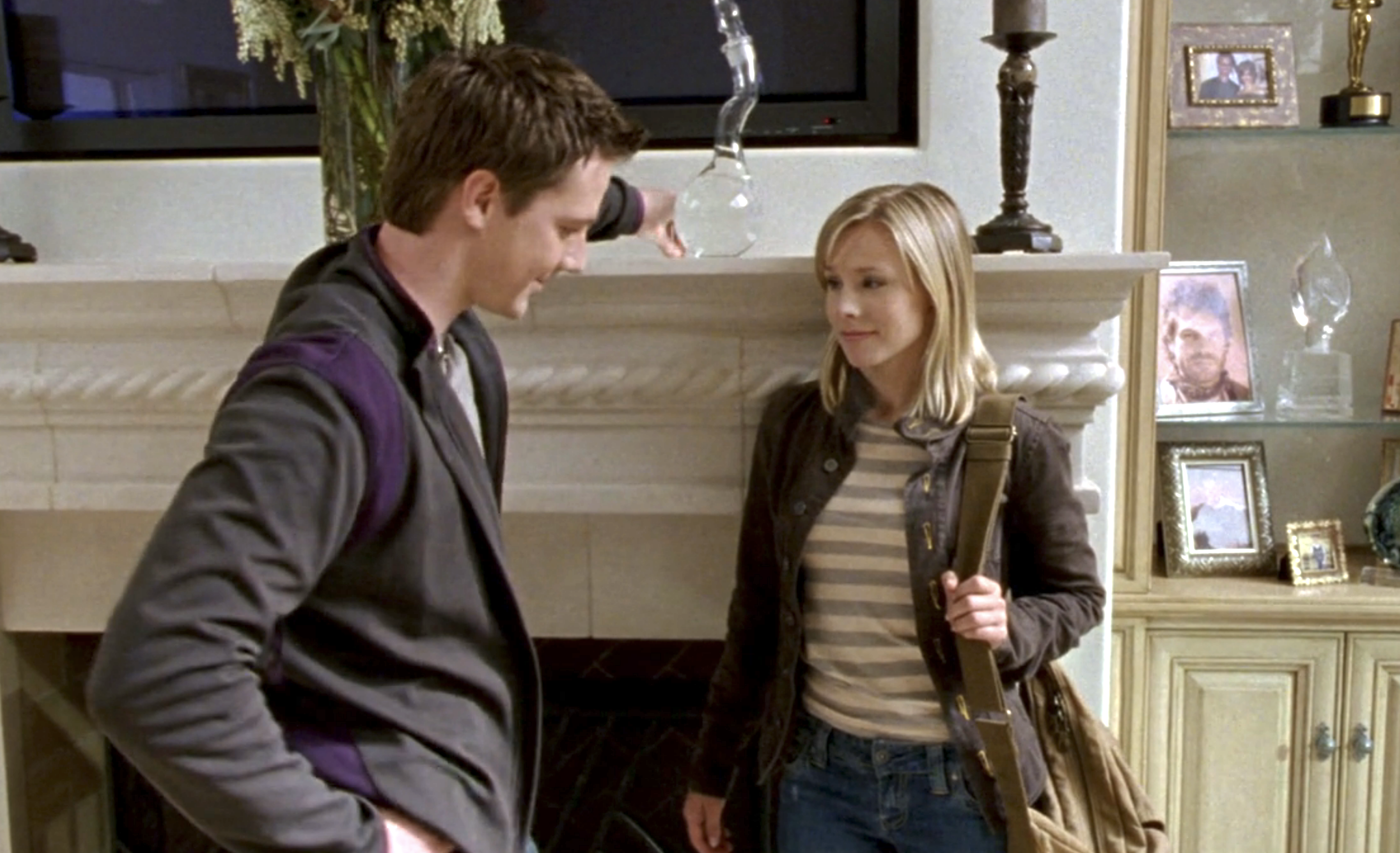 Screenshot from S1E20 of Veronica Mars. Logan is leaning on the mantle in the living room smiling. Veronica has her back to the mantle is smiling sheepishly.