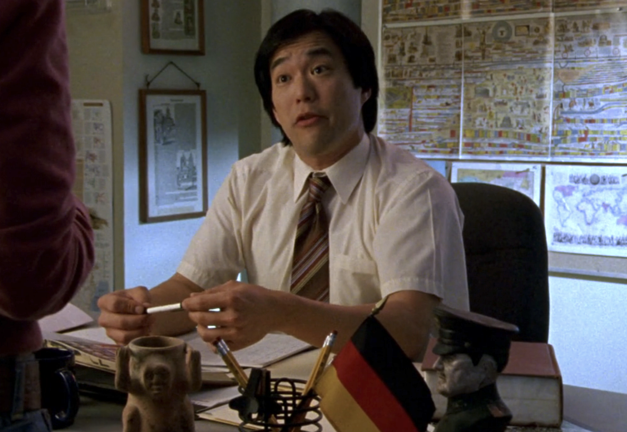 Screenshot of S1E20 of Veronica Mars. Mr. Wu is sitting at the head of the class at a teacher's desk talking to Veronica who is off screen. Sitting on the desk is a German flag and a small busy of a soldier.
