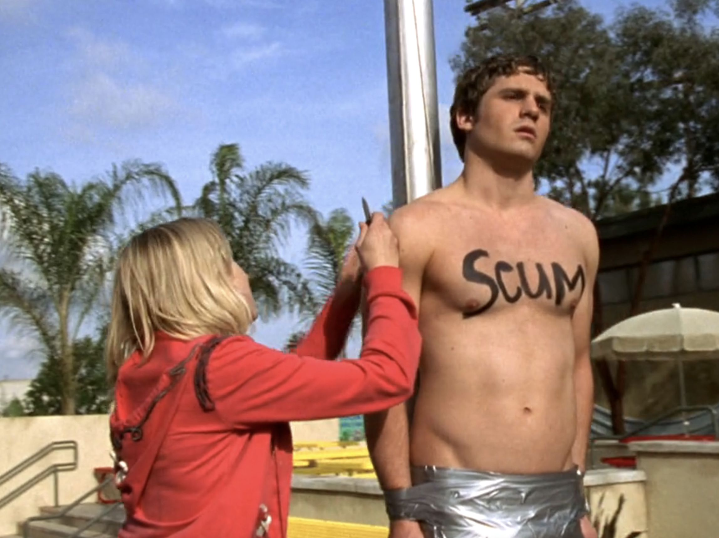 Screenshot from S1E20 of veronica Mars. Tad is taped to the flagpole naked. "SCUM" is written on his chest in black. Veronica is about to cut him down from the flagpole with a knife.