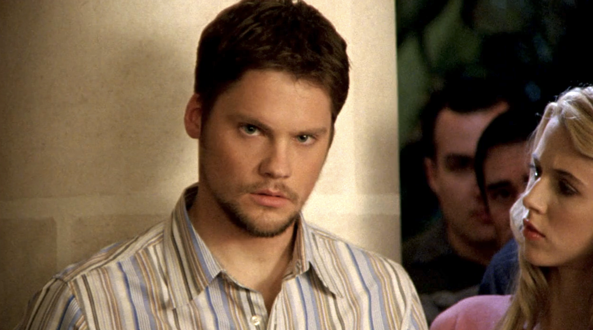 Screenshot from S1E21 of Veronica Mars. Duncan is looking upset at something offscreen. He's wearing a striped button down shirt. Meg is standing to his left looking at him.