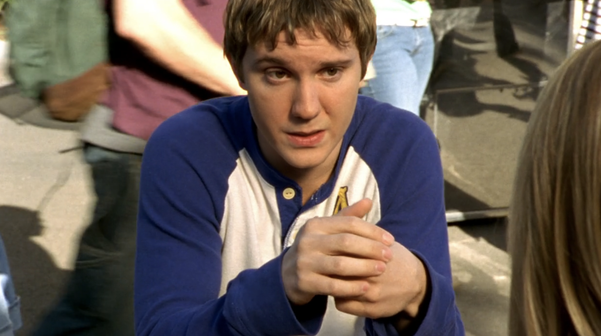 Screenshot from S1E21 of Veronica Mars. Luke is wearing a baseball shirt with blue sleeves looking at an offscreen Veronica. He's sitting at an outdoor lunch table. His hands are clasped in front of him and his elbows are on the table.