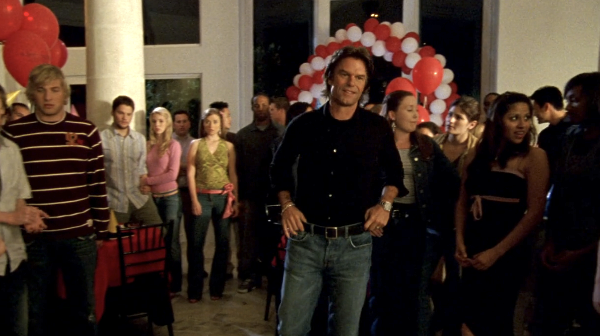 Screnshot from S1E21 of Veronica Mars. Aaron Echools is standing in his house which is crowded with Logan's friends. His hands are on his hips and he's smiling slightly.