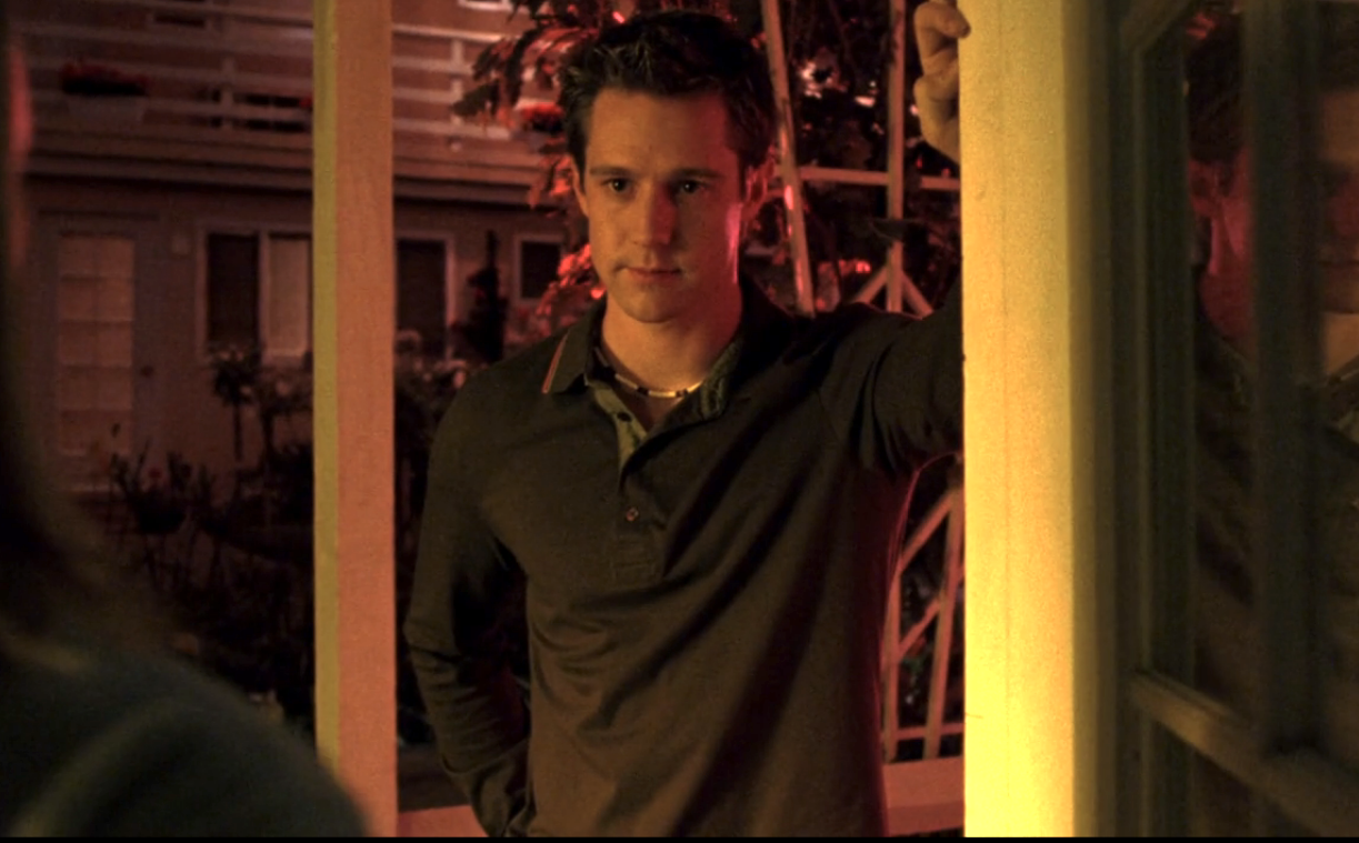 Screenshot from S1E21 of Veronica Mars. Logan is standing outside in a brown shirt and puka shell necklace. He is leaning on a door frame. He looks serious.