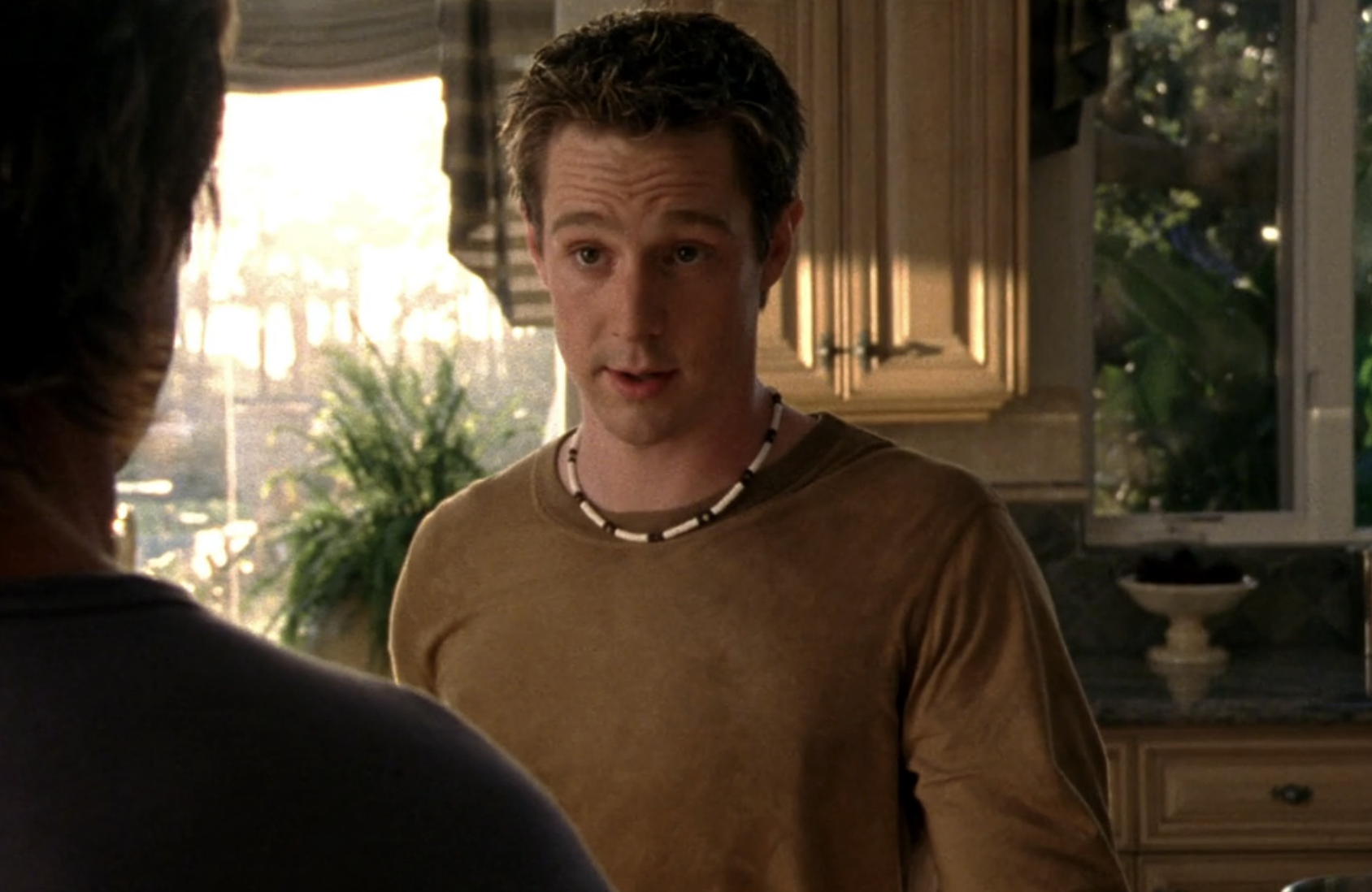 Screenshot from S1E21 of Veronica Mars. Logan is in his kitchen, wearing a brown long-sleeve tee and puka shell necklass. His eyebrows are rasied, he's in converastion with Aaron, whose back is to us.